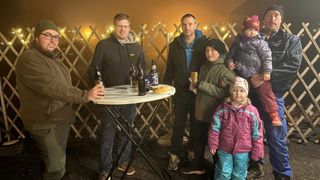 Eine Gruppe von Menschen steht lächelnd für ein Foto zusammen an einem Tisch. Sie befinden sich in einer festlichen Atmosphäre mit Lichtern und Dekorationen im Hintergrund. Auf dem Tisch stehen Flaschen und Essen.