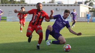 Zwei Fußballspieler konkurrieren auf einem Feld. Einer trägt Rot, der andere Lila. Der Rote läuft, der Lila kniet, beide konzentrieren sich auf den Ball.