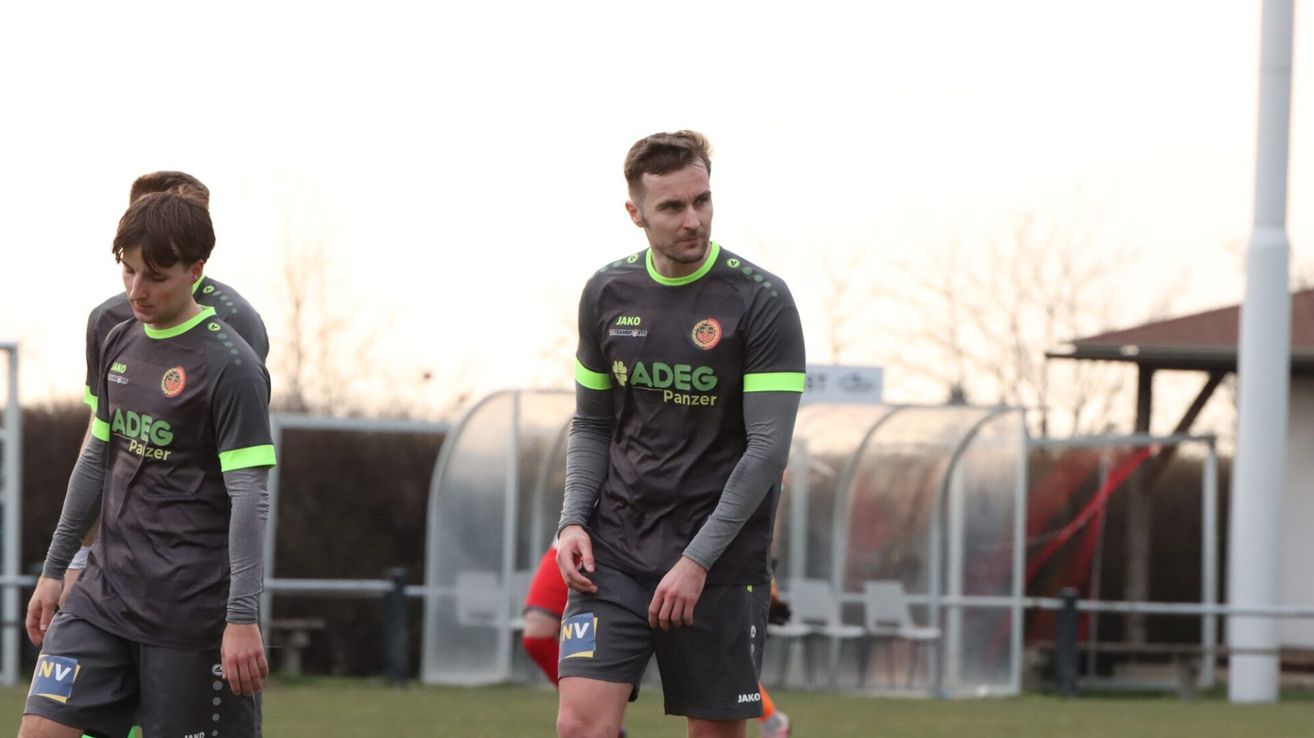 Ein Fußballspieler in einem schwarzen und grünen Trikot mit den Buchstaben ADEG auf der Vorderseite, steht auf einem Fußballfeld.