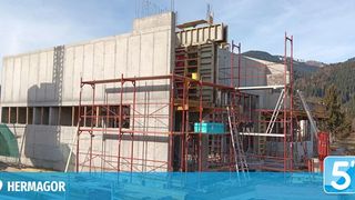 Ein Betongebäude im Bau mit roten Gerüsten. Im Hintergrund sind Berge sichtbar, und der Standort befindet sich in der Nähe eines Ortes namens Hermagor.