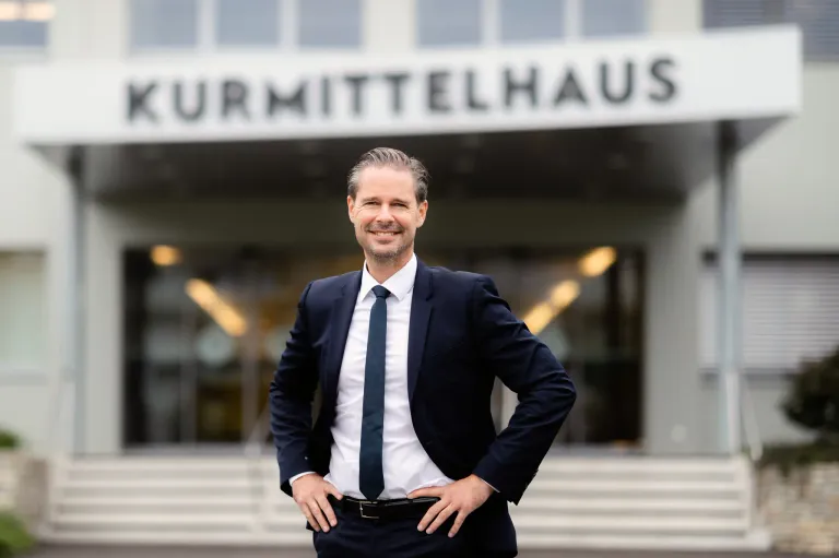 Ein Mann in einem Anzug steht lächelnd mit den Händen auf den Hüften vor dem Kurmittelhaus. Das Gebäude hat Glasfenster und ein weißes Schild darüber.