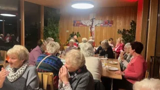 Eine Gruppe älterer Frauen sitzt um einen Esstisch in einem Raum herum, möglicherweise bei einer religiösen Zusammenkunft. Einige beten, während andere in Gespräche vertieft sind. Ein Kreuz ist an der Wand über ihnen angebracht.