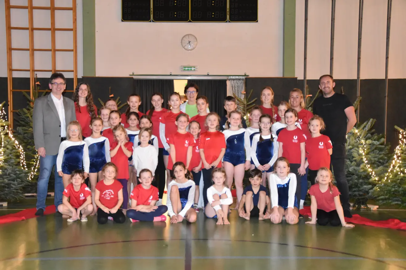 Eine Gruppe von Kindern und Erwachsenen in roten und blauen Uniformen posiert für ein Foto auf einem Spielfeld. Im Hintergrund befinden sich Uhren und eine Uhr an der Wand.
