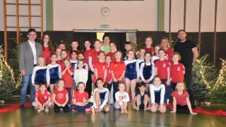 Eine Gruppe von Kindern und Erwachsenen in roten und blauen Uniformen posiert für ein Foto auf einem Spielfeld. Im Hintergrund befinden sich Uhren und eine Uhr an der Wand.