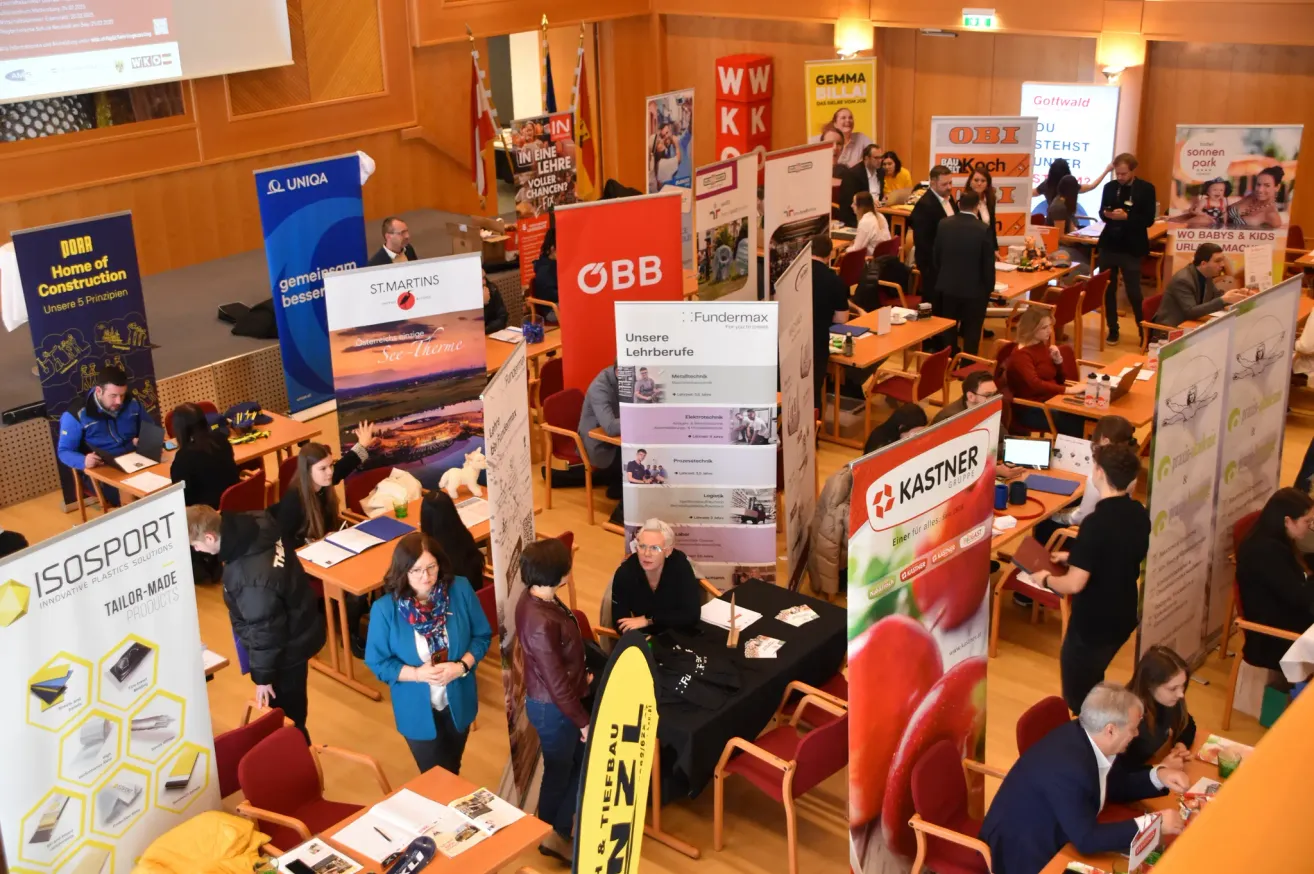 Eine Indoor-Veranstaltung mit verschiedenen Bannern und Tischen. Die Leute sitzen an Tischen, möglicherweise bei einer Jobmesse. Eine Person steht in der Nähe eines Standes mit einem 'Fundermax'-Banner. Eine Gruppe von Menschen unterhält sich in der Nähe eines Standes mit einem 'Kastner'-Banner.