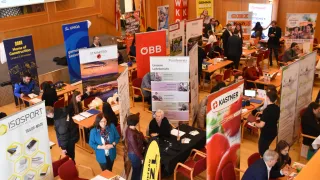 Eine Indoor-Veranstaltung mit verschiedenen Bannern und Tischen. Die Leute sitzen an Tischen, möglicherweise bei einer Jobmesse. Eine Person steht in der Nähe eines Standes mit einem 'Fundermax'-Banner. Eine Gruppe von Menschen unterhält sich in der Nähe eines Standes mit einem 'Kastner'-Banner.