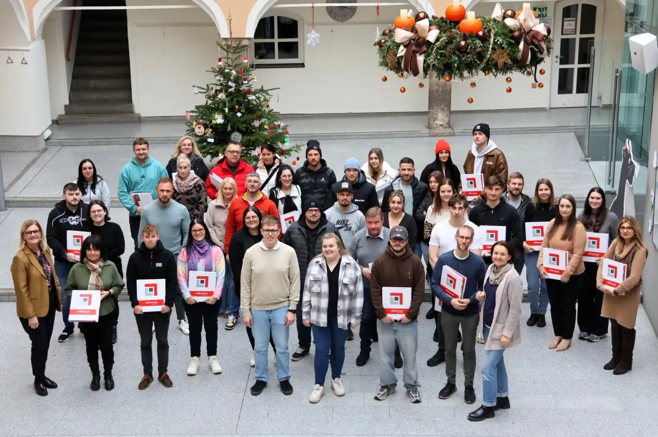 Eine Gruppe von Menschen steht auf einem Platz mit einem Weihnachtsbaum und posiert für ein Foto. Einige halten Zertifikate, und es gibt dekorative Ornamente darüber.