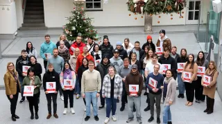 Eine Gruppe von Menschen steht auf einem Platz mit einem Weihnachtsbaum und posiert für ein Foto. Einige halten Zertifikate, und es gibt dekorative Ornamente darüber.
