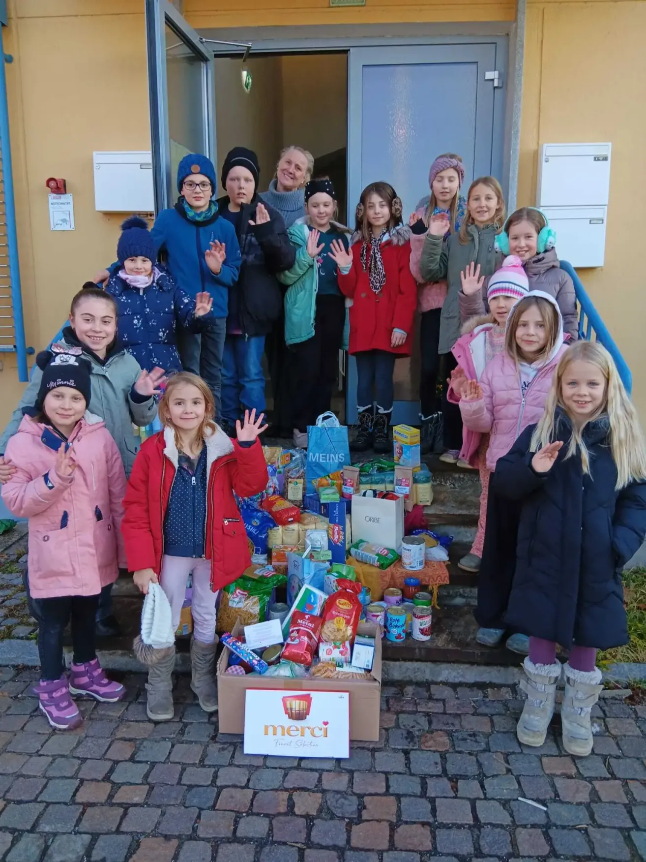 Eine Gruppe von Kindern und Erwachsenen, alle in Winterkleidung, steht auf den Stufen vor einem Gebäude. Sie lächeln und machen Friedenszeichen. Vor ihnen stehen Kisten und Säcke mit Lebensmitteln. Die Kisten haben das Wort 'Merci' darauf.