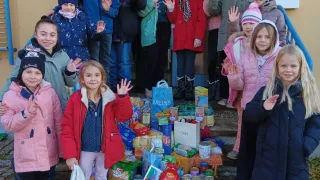 Eine Gruppe von Kindern und Erwachsenen, alle in Winterkleidung, steht auf den Stufen vor einem Gebäude. Sie lächeln und machen Friedenszeichen. Vor ihnen stehen Kisten und Säcke mit Lebensmitteln. Die Kisten haben das Wort 'Merci' darauf.