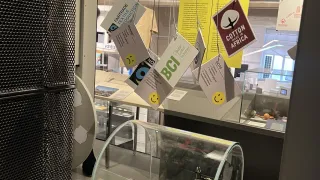 Eine Museumsausstellung mit einem Glaskasten, der eine alte Nähmaschine und eine grüne Box enthält. Mehrere hängende Schilder mit Text sind sichtbar, darunter eines mit der Aufschrift 'Fairtrade' und ein anderes mit der Aufschrift 'Baumwolle hergestellt in Afrika'.