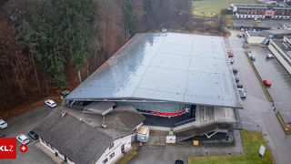 Luftaufnahme eines großen Gebäudes mit Metalldach und rot-weißer Wand. Bäume und ein Rasen umgeben das Gebäude. Autos sind auf der Straße geparkt.