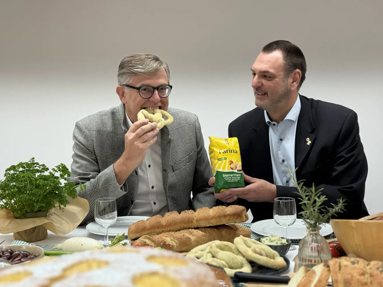 Zwei Männer sitzen an einem Tisch, einer isst ein Brezel, während der andere eine Tüte Brezeln hält. Der Tisch ist mit verschiedenen Speisen beladen, darunter Brot, und zwei Weingläsern.