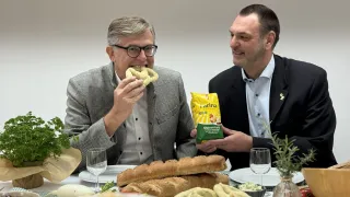 Zwei Männer sitzen an einem Tisch, einer isst ein Brezel, während der andere eine Tüte Brezeln hält. Der Tisch ist mit verschiedenen Speisen beladen, darunter Brot, und zwei Weingläsern.