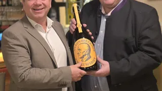 Zwei Männer halten eine Bierflasche mit einem lächelnden Gesicht und stehen in einer Bar. Hinter ihnen stehen Regale mit Flaschen und Gläsern. Ein gerahmtes Bild ist an der Wand angebracht.