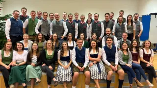 Eine Gruppe von Menschen in traditioneller bayerischer Kleidung posiert für ein Gruppenfoto in einem Raum. Die Gruppe besteht aus Männern und Frauen, und sie lächeln und posieren für die Kamera. Einige von ihnen tragen Brillen, und einige tragen Lederschuhe. Der Raum hat weiße Wände und eine Holzbank.