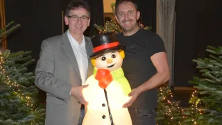 Zwei Männer in festlicher Kleidung stehen neben einem großen beleuchteten Schneemann und lächeln für die Kamera vor einem geschmückten Weihnachtsbaum.