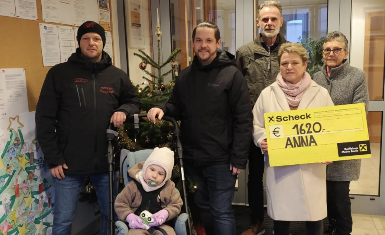 Eine Familie steht vor einem geschmückten Weihnachtsbaum. Ein Mann in einem schwarzen Mantel hält einen Kinderwagen mit einem Kind in einer Wintermütze. Daneben hält eine Frau ein Schild mit den Worten 'x Check' und '162 Anna'.