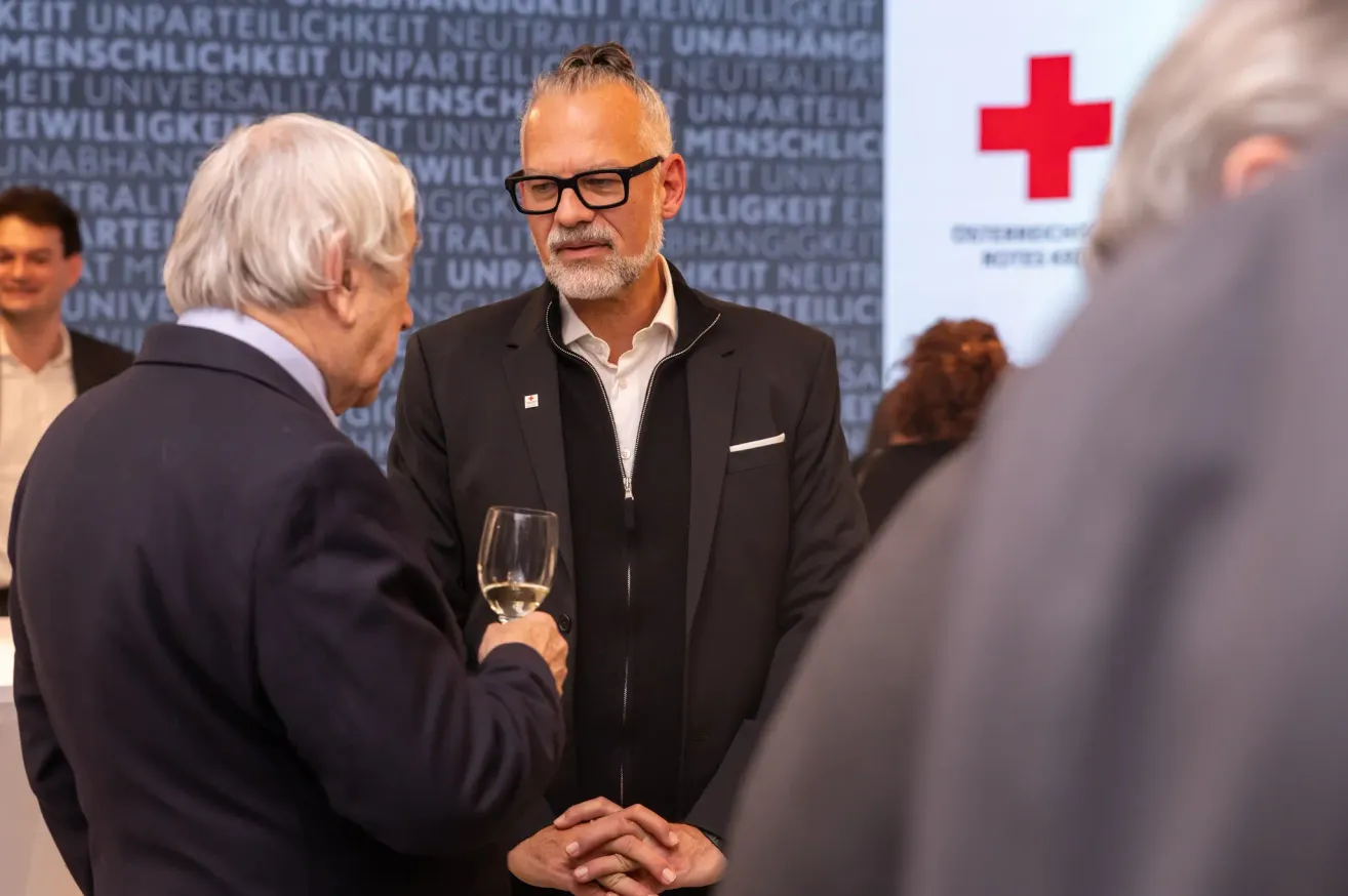 Zwei Männer in Anzügen unterhalten sich; einer hält ein Weinglas. Sie stehen vor einer Wand mit Wörtern in einer fremden Sprache und einem roten Kreuz-Symbol.