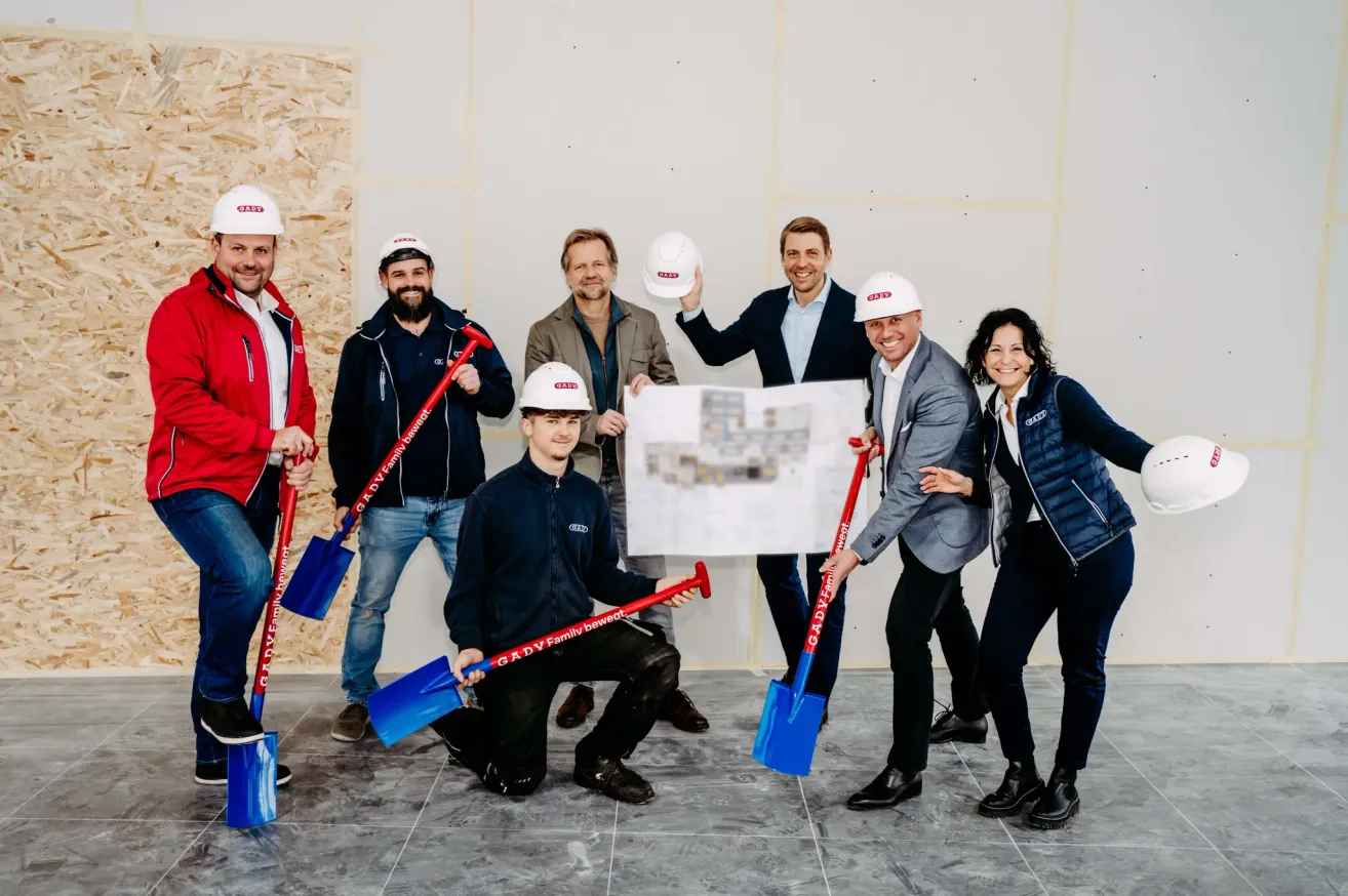 Eine Gruppe von Menschen in Jacken und weißen Helmen posiert mit Schaufeln in einem Raum. Ein Mann in der Mitte hält ein Stück Papier.