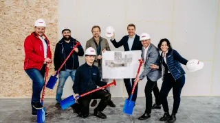 Eine Gruppe von Menschen in Jacken und weißen Helmen posiert mit Schaufeln in einem Raum. Ein Mann in der Mitte hält ein Stück Papier.