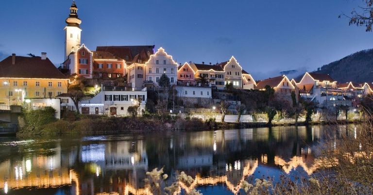 Eine Stadt mit beleuchteten Gebäuden wird bei Nacht gezeigt, mit Blick auf einen Fluss. Die Stadt hat eine Mischung aus Architekturstilen, wobei einige Gebäude Türme und andere moderne Designs aufweisen.
