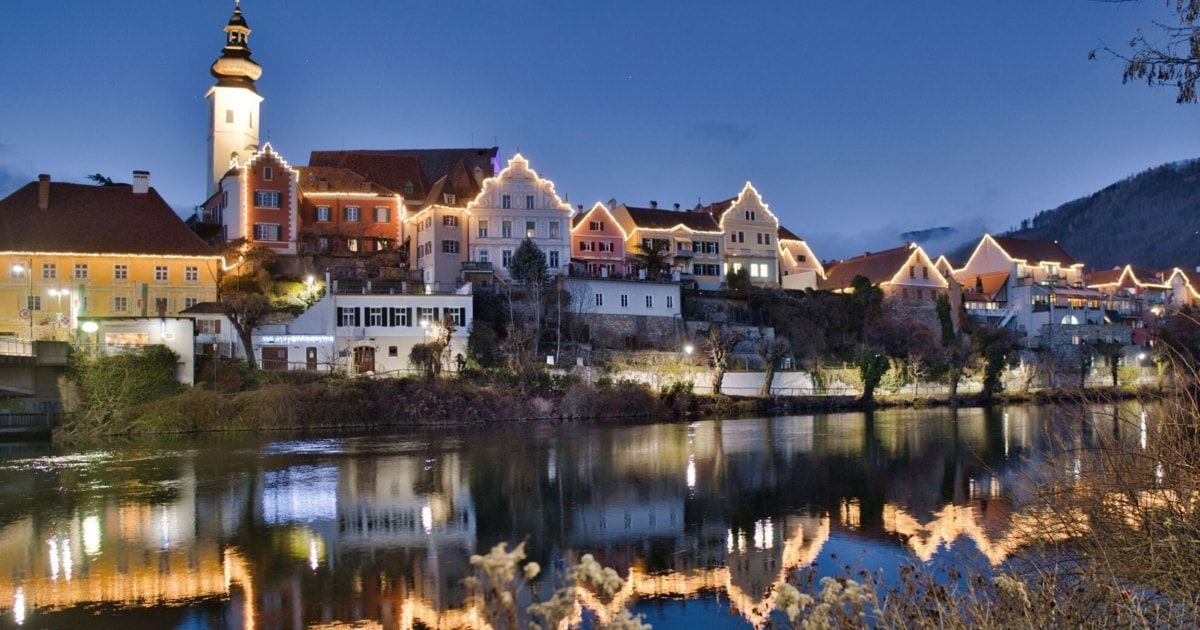 Eine Stadt mit beleuchteten Gebäuden wird bei Nacht gezeigt, mit Blick auf einen Fluss. Die Stadt hat eine Mischung aus Architekturstilen, wobei einige Gebäude Türme und andere moderne Designs aufweisen.