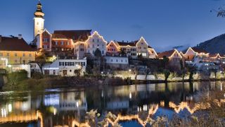 Eine Stadt mit beleuchteten Gebäuden wird bei Nacht gezeigt, mit Blick auf einen Fluss. Die Stadt hat eine Mischung aus Architekturstilen, wobei einige Gebäude Türme und andere moderne Designs aufweisen.