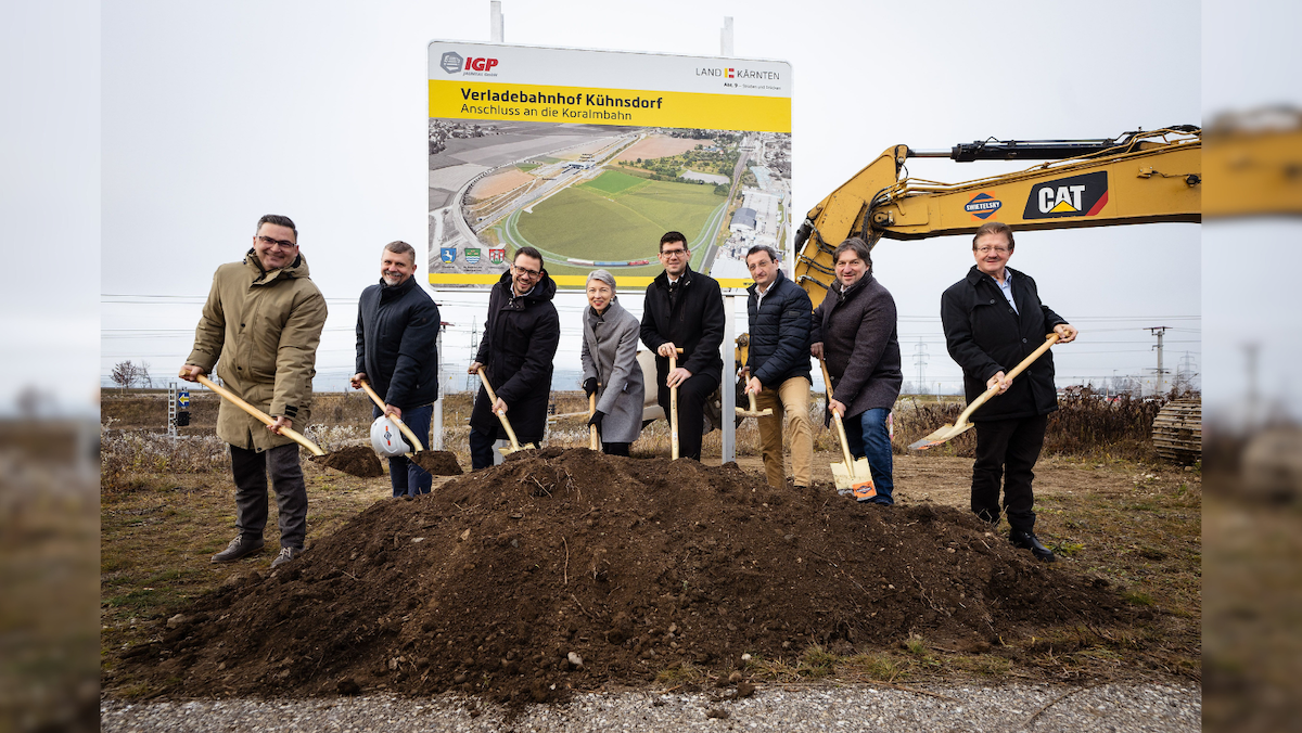 Eine Gruppe von Menschen versammelt sich für eine Spatenstich-Zeremonie mit Schaufeln. Sie stehen vor einem Schild und einem gelben Bagger. Das Schild zeigt 'Verladebahnhof Kühnsdorf Anschluß an die Koralmbahn.' Die Gruppe lächelt und posiert für ein Foto.