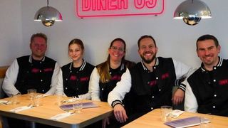 Fünf Menschen posieren für ein Foto in einem Restaurant. Sie tragen schwarze und weiße Jacken. Der Name des Restaurants lautet Diner 69.