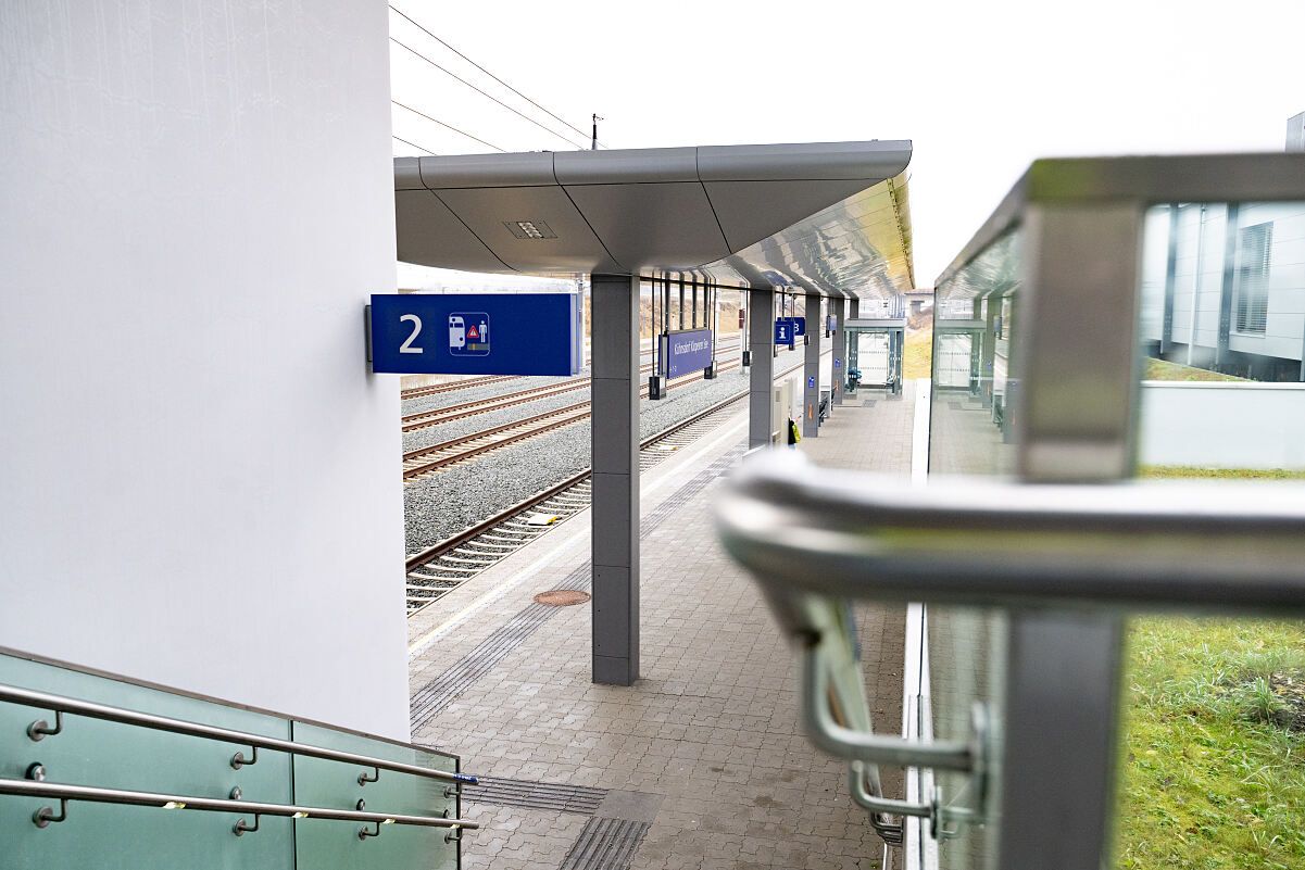 Ein moderner Bahnhof mit einer Plattform mit der Nummer 2, gekennzeichnet durch ein blaues Schild mit einer Person und einem Rollstuhlsymbol, einem Überdach und einem klaren Blick auf die Gleise.