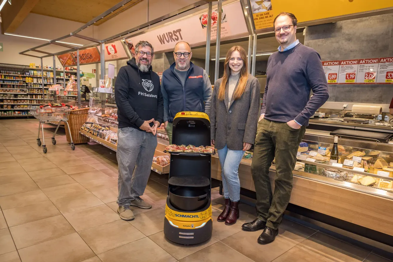 Vier Personen stehen in einem Lebensmittelgeschäft und lächeln für ein Foto. Eine Frau hält eine Pizza in einem Roboter. Hinter ihnen befindet sich eine Fleischtheke.