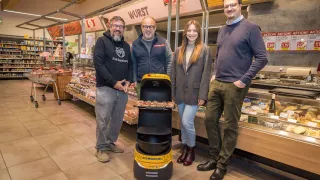 Vier Personen stehen in einem Lebensmittelgeschäft und lächeln für ein Foto. Eine Frau hält eine Pizza in einem Roboter. Hinter ihnen befindet sich eine Fleischtheke.