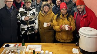 Eine Gruppe von Menschen lächelt und posiert für ein Foto. Ein Mann in Tarnkleidung hält einen Becher. Zwei Frauen in gelben Mänteln halten Tassen. Eine Frau in einer roten Mütze trägt einen Ring. Flaschen und Servietten liegen auf einem Tisch.