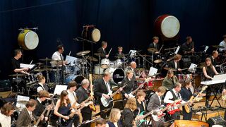 Eine Gruppe von Musikern spielt auf der Bühne mit verschiedenen Instrumenten, einschließlich Trommeln, Gitarren und Becken, vor einem blauen Hintergrund.