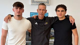 Drei Männer stehen zusammen drinnen, einer in einem weißen Pullover, ein anderer in einer schwarzen adidas-Kapuzenjacke und der dritte in einem schwarzen T-Shirt. Hinter ihnen befindet sich eine Wand mit einem blauen Schild und einem silbernen Rahmen.