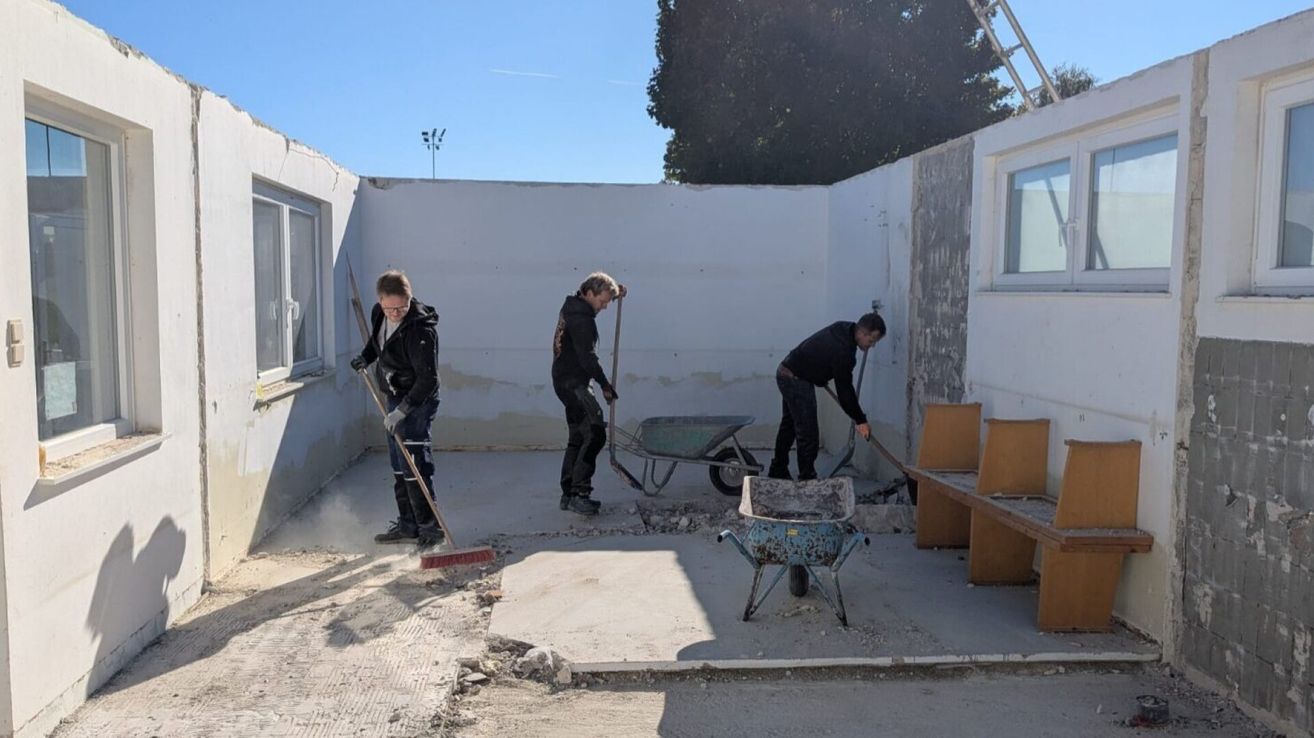 Drei Arbeiter reinigen einen Außenbereich. Einer fegt, ein anderer benutzt eine Schubkarre und der dritte arbeitet an der Wand. Sie tragen Handschuhe und schwarze Jacken.