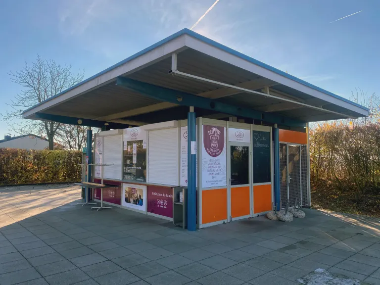 Ein kleiner orangefarbener und weißer Kiosk mit einem Dach steht an der Straßenseite. Es gibt zwei Tische mit Stühlen draußen. Der Kiosk hat eine Glastür und ein Glasfenster.