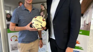 Zwei Männer stehen nebeneinander, einer hält einen Fußball. Sie stehen vor einem grünen Banner mit Logos und Text.