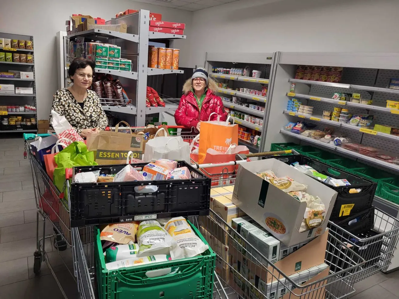 Zwei Frauen stehen in einem Lebensmittelgeschäft mit vollbepackten Einkaufswagen. Eine trägt ein Leopardenmuster-Shirt, die andere eine rote Jacke. Die Regale hinter ihnen sind mit verschiedenen Produkten bestückt.