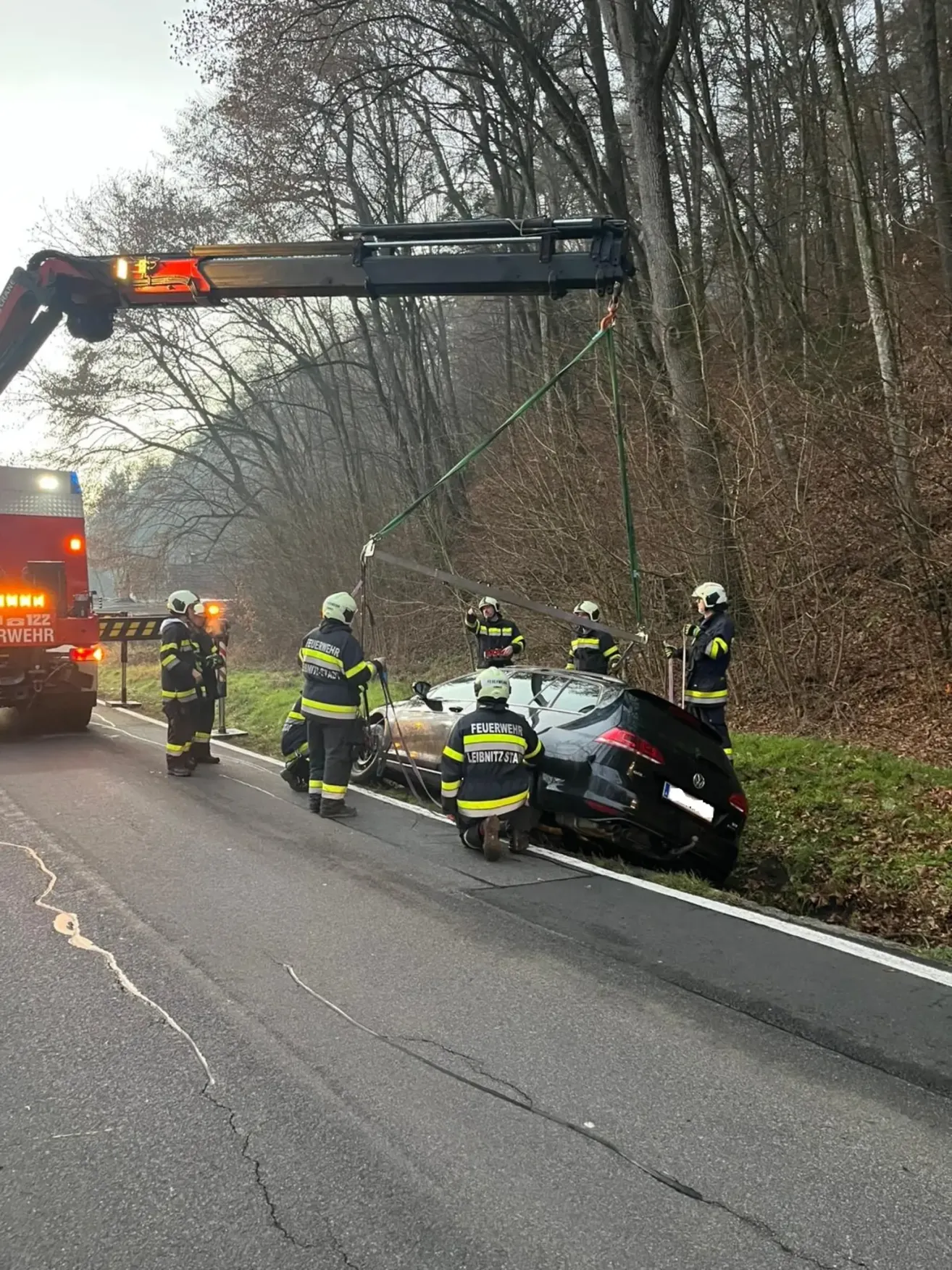 Feuerwehrleute entfernen ein verunglücktes schwarzes Auto mit einem Kran. Die Szene spielt am Straßenrand mit Bäumen und einem roten Notfallfahrzeug im Hintergrund.
