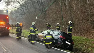 Feuerwehrleute entfernen ein verunglücktes schwarzes Auto mit einem Kran. Die Szene spielt am Straßenrand mit Bäumen und einem roten Notfallfahrzeug im Hintergrund.