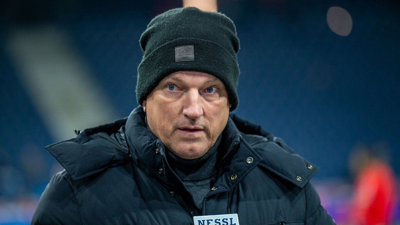 Ein Mann in einem dunkelblauen Mantel und einer schwarzen Mütze mit Logo, der ernsthaft dreinblickt. Vor ihm ist ein Mikrofon mit der Aufschrift NESSL zu sehen.