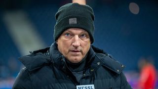 Ein Mann in einem dunkelblauen Mantel und einer schwarzen Mütze mit Logo, der ernsthaft dreinblickt. Vor ihm ist ein Mikrofon mit der Aufschrift NESSL zu sehen.