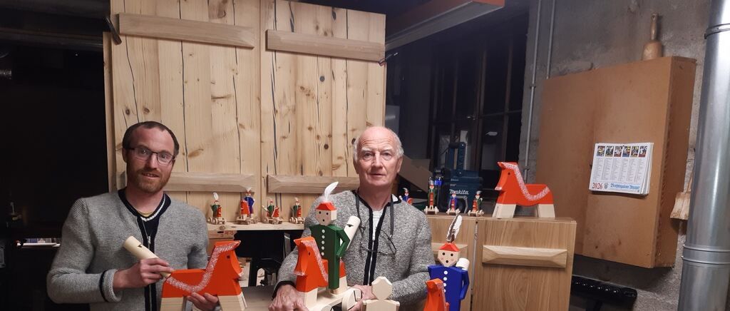 Zwei Männer posieren mit Holzspielzeug in einer Werkstatt, einer hält ein Spielzeugpferd, der andere eine Spielzeugfigur. Hinter ihnen befinden sich Holzregale und Türen.