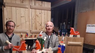 Zwei Männer posieren mit Holzspielzeug in einer Werkstatt, einer hält ein Spielzeugpferd, der andere eine Spielzeugfigur. Hinter ihnen befinden sich Holzregale und Türen.