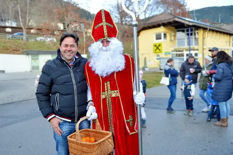 Ein Mann in der Kleidung von Sankt Nikolaus mit einem Korb voller Orangen steht neben einem lächelnden Mann in einer Jacke an einem sonnigen Tag.