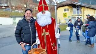 Ein Mann in der Kleidung von Sankt Nikolaus mit einem Korb voller Orangen steht neben einem lächelnden Mann in einer Jacke an einem sonnigen Tag.