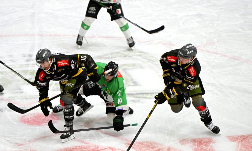 Eishockeyspieler konkurrieren auf einer Eisbahn. Ein Spieler stürzt, während ein anderer versucht, den Puck zu bekommen. Beide Spieler tragen Helme und Handschuhe.