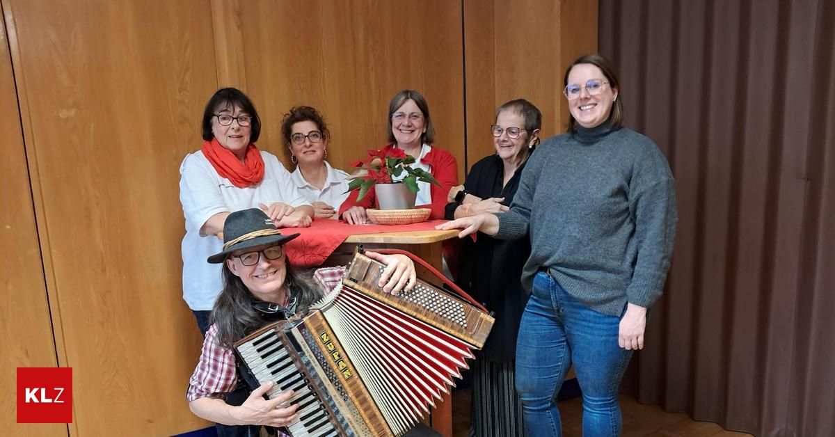 Eine Gruppe lächelnder Frauen und ein Mann mit einem Akkordeon stehen hinter einem Tisch mit einem Topf auf ihm.